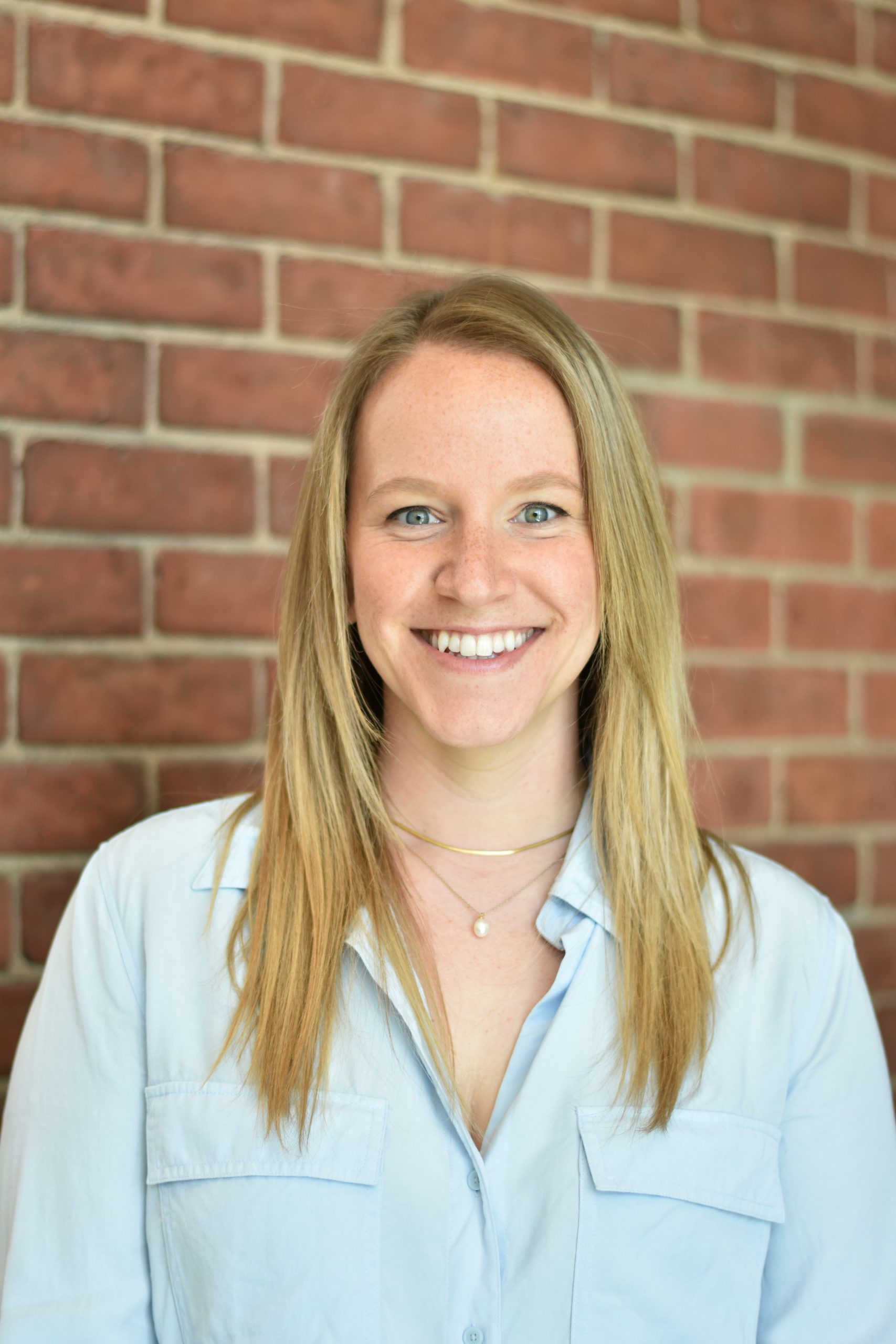 A photo of Janet Lawson. Janet has long blonde hair and a light blue button down shirt. She is smiling and standing against a red brick background.